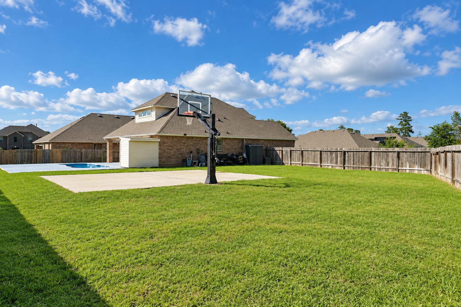 A spacious backyard with a basketball hoop, swimming pool and lawn behind a brick home